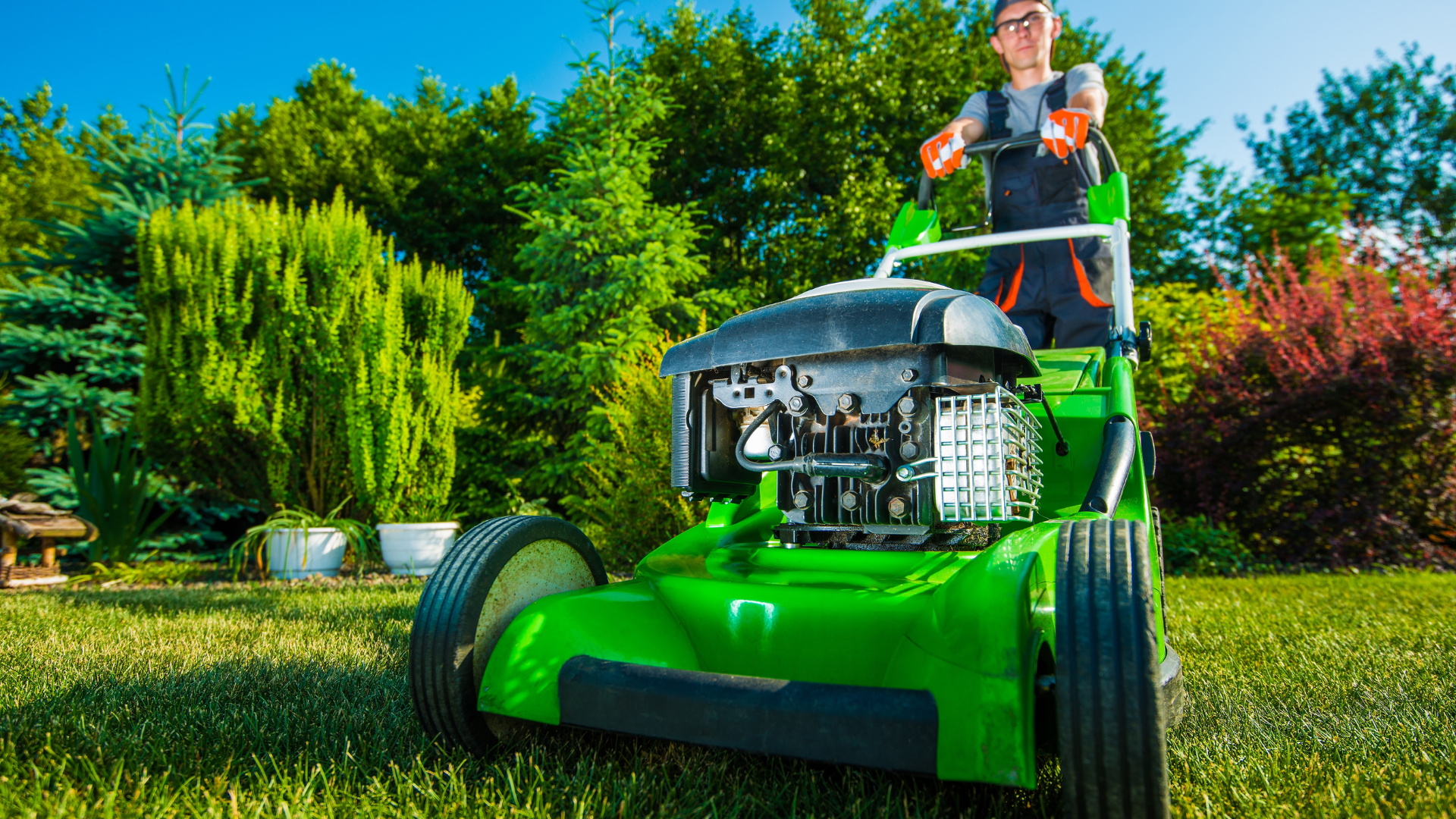 Person mowing a green lawn with a green lawnmower in a sunny backyard with trees.