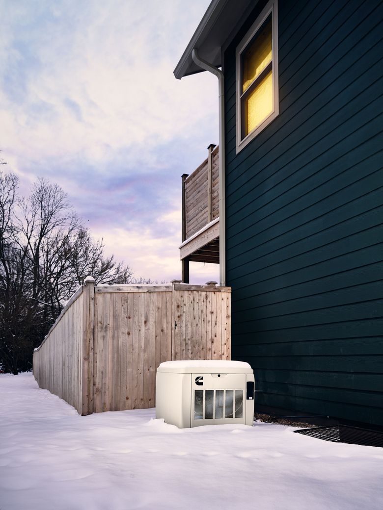 Snowy backyard beside a dark green house with a lit window, wooden fence, and outdoor AC unit.