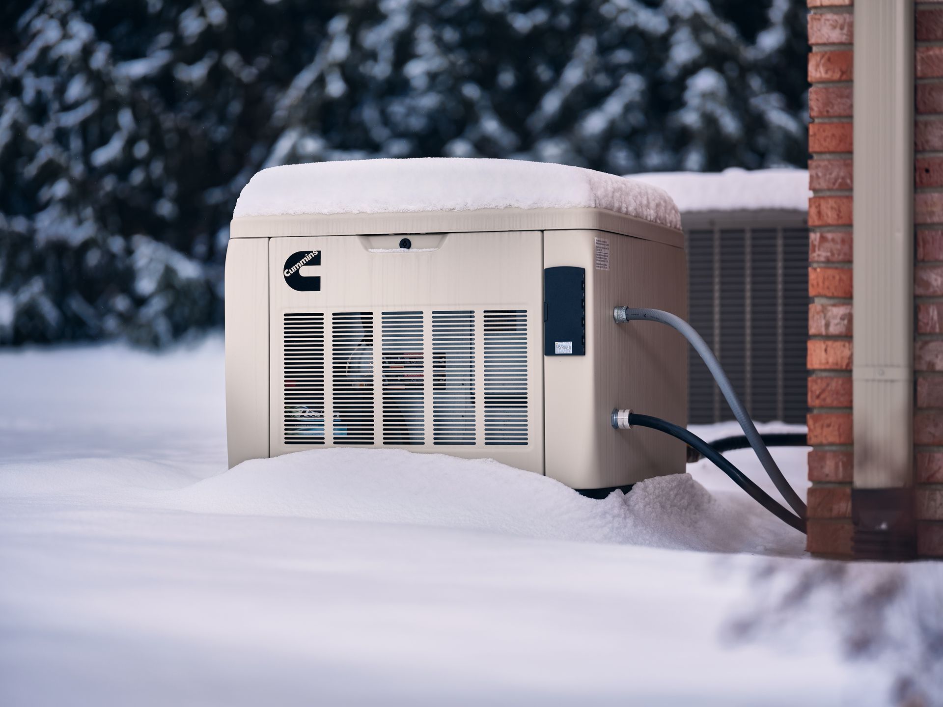 Outdoor air-conditioning unit buried in snow beside a brick wall, with visible pipes and snowy trees in the background