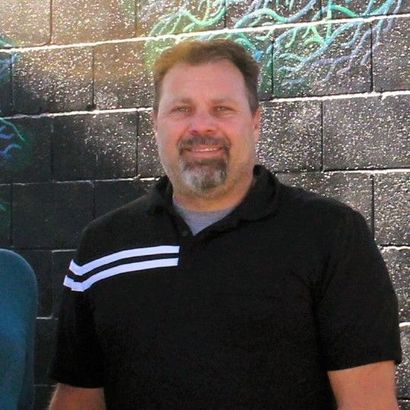 Smiling man in a black polo shirt with white stripes standing against a dark brick wall