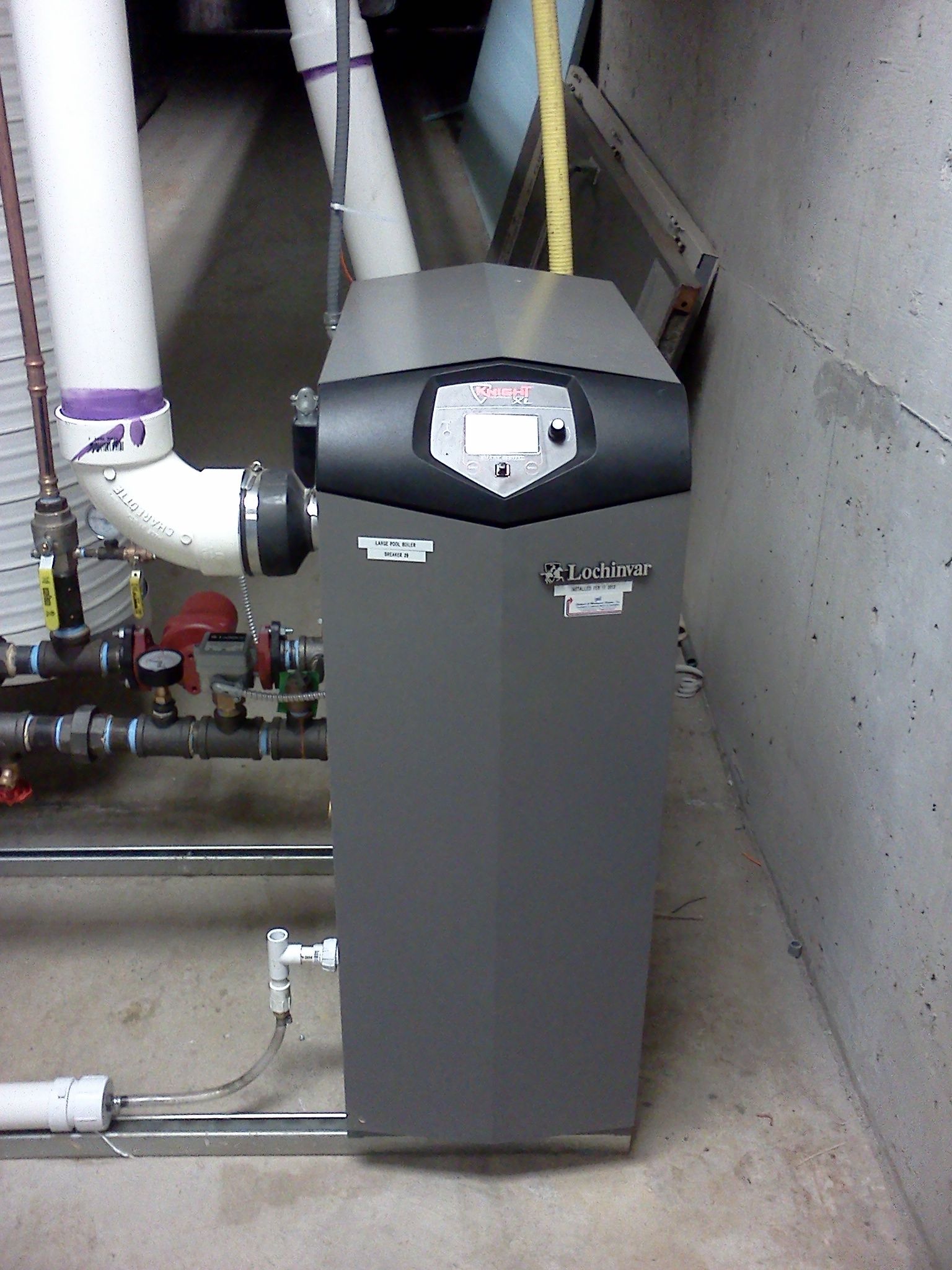 Gray furnace in a utility room with white pipes and ductwork against concrete walls.