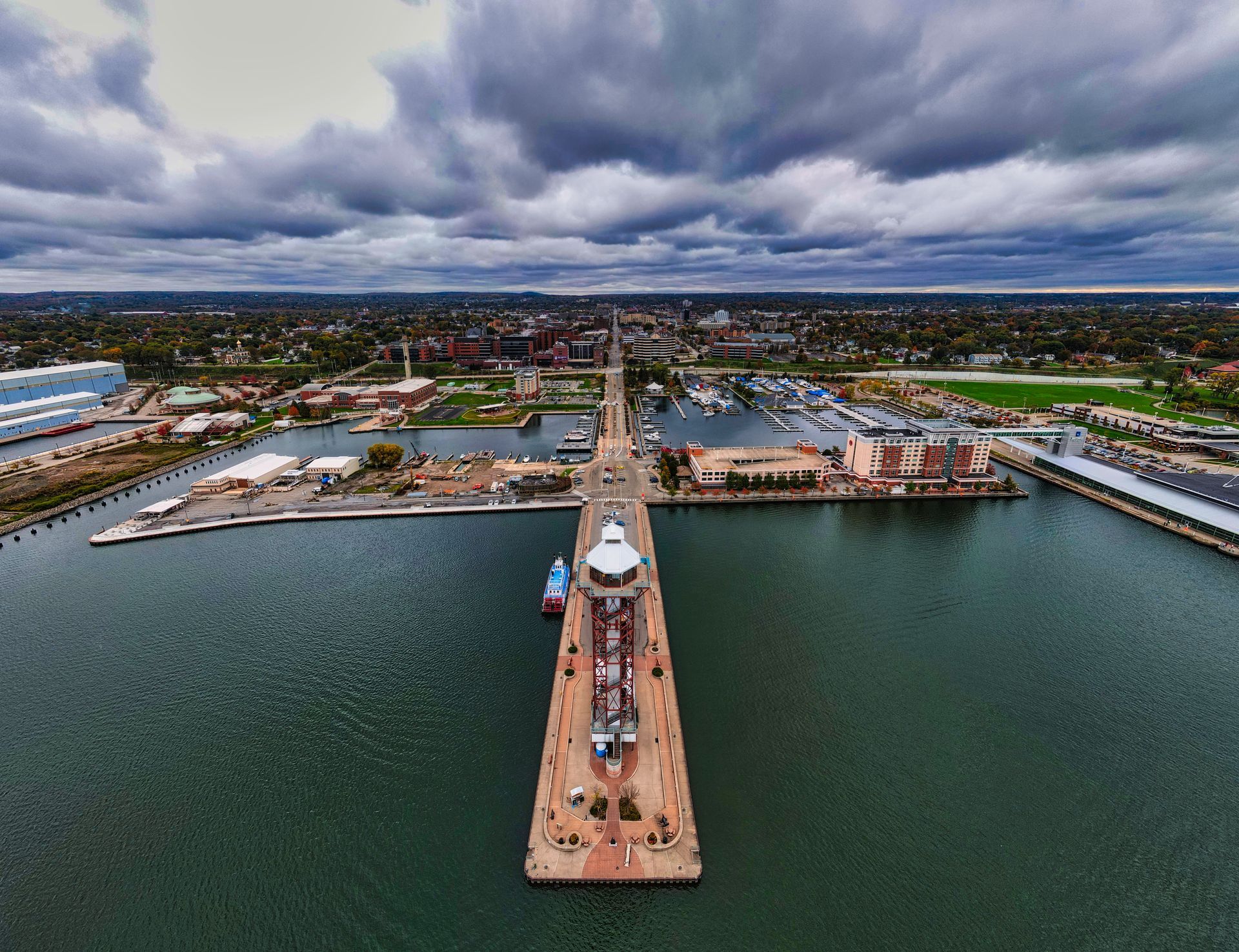 Aerial view of a marina pier extending into dark water under cloudy skies, with buildings on both sides.