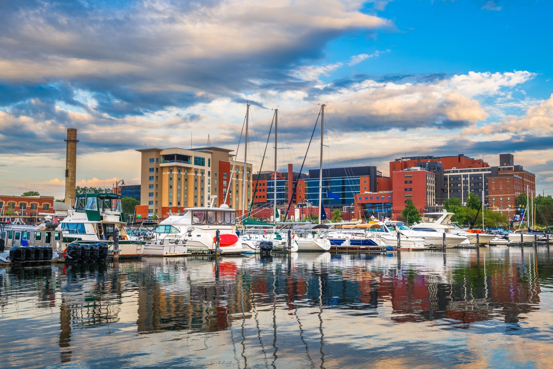 Marina with white sailboats, harbor buildings, and cloudy sky reflected in calm water
