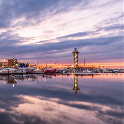 Sunset over a waterfront harbor with a lit tower and boats reflected in calm water