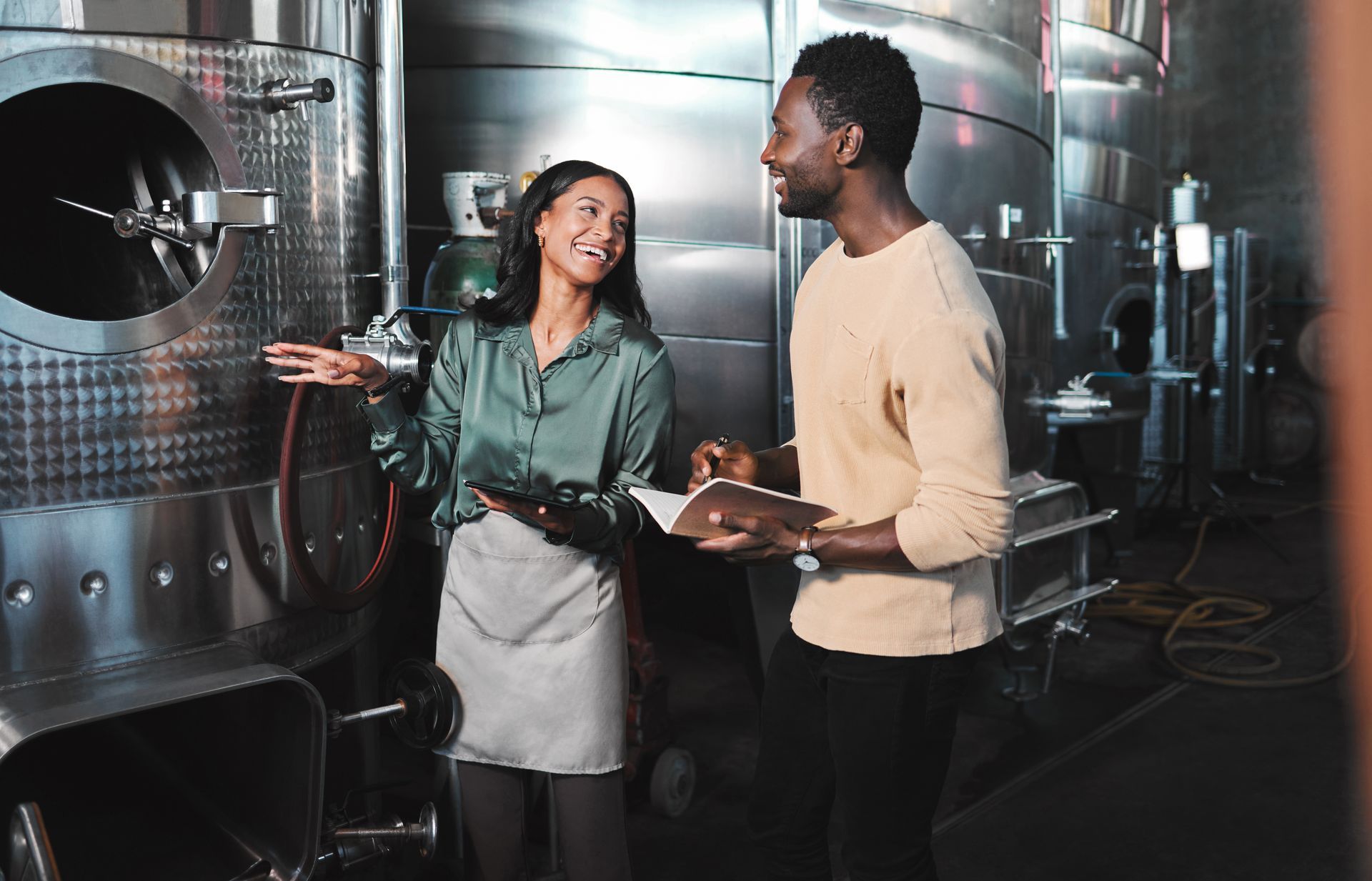 Two coworkers in a brewery discussing beside large stainless steel tanks