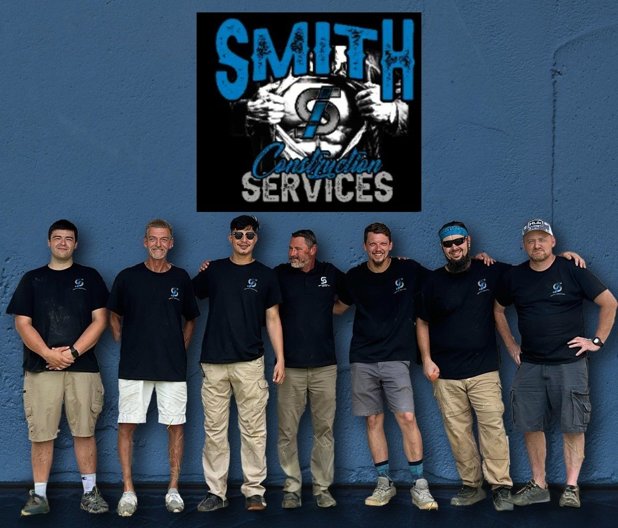 Team of seven men in black shirts and khaki pants pose in front of a blue wall below a logo.