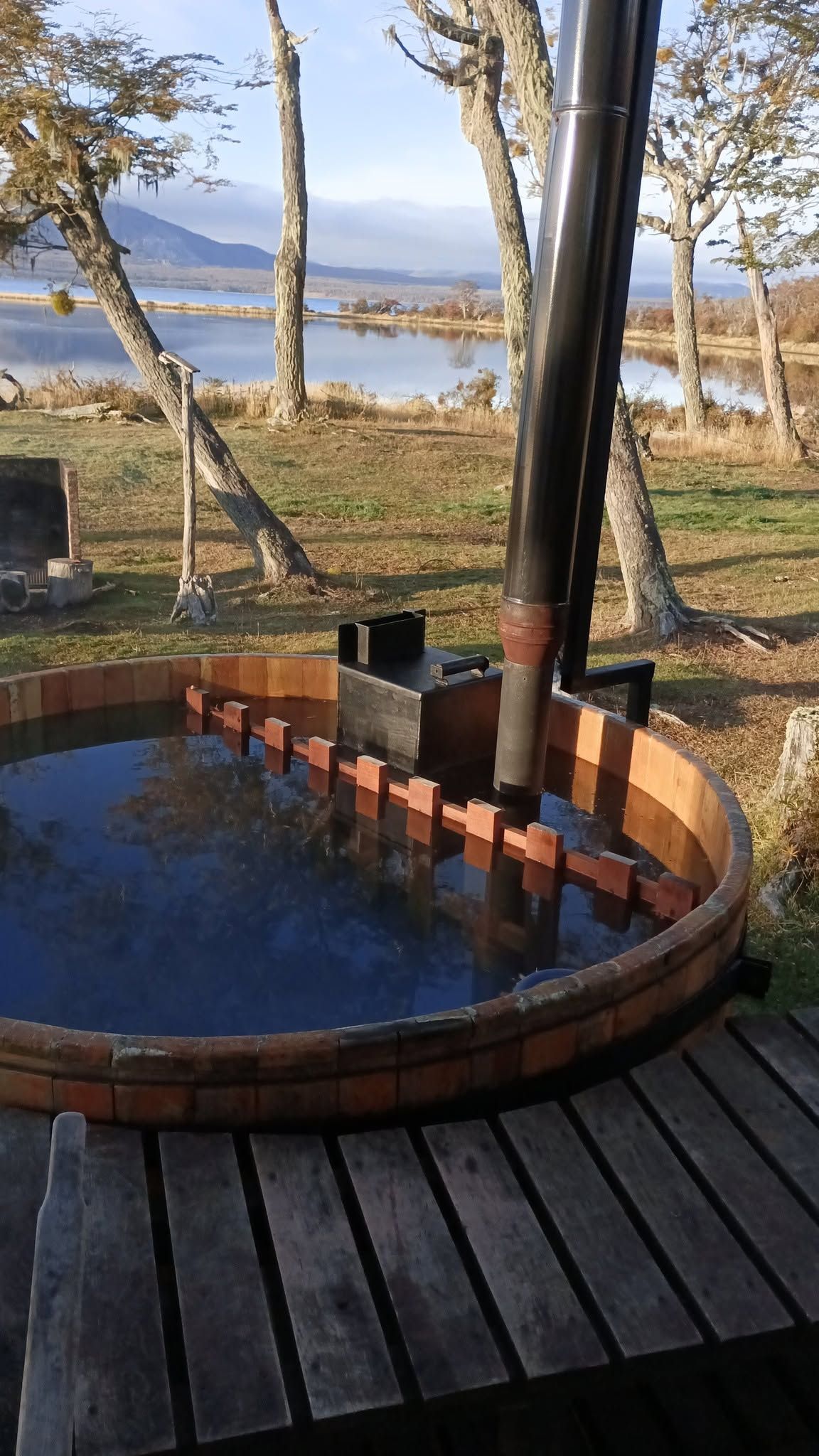 Bañera de hidromasaje exterior de madera con agua oscura, con vistas a un lago y árboles en una terraza.