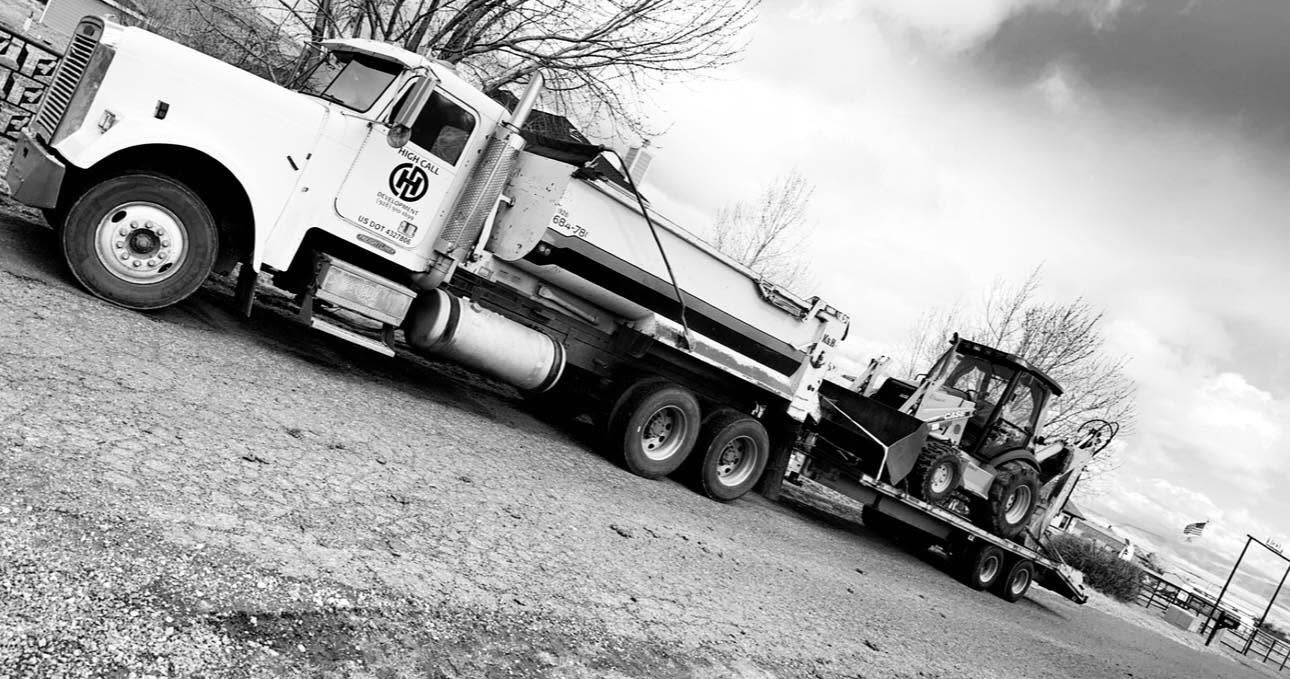 Tilted black-and-white photo of a flatbed truck hauling a bulldozer on a gravel lot