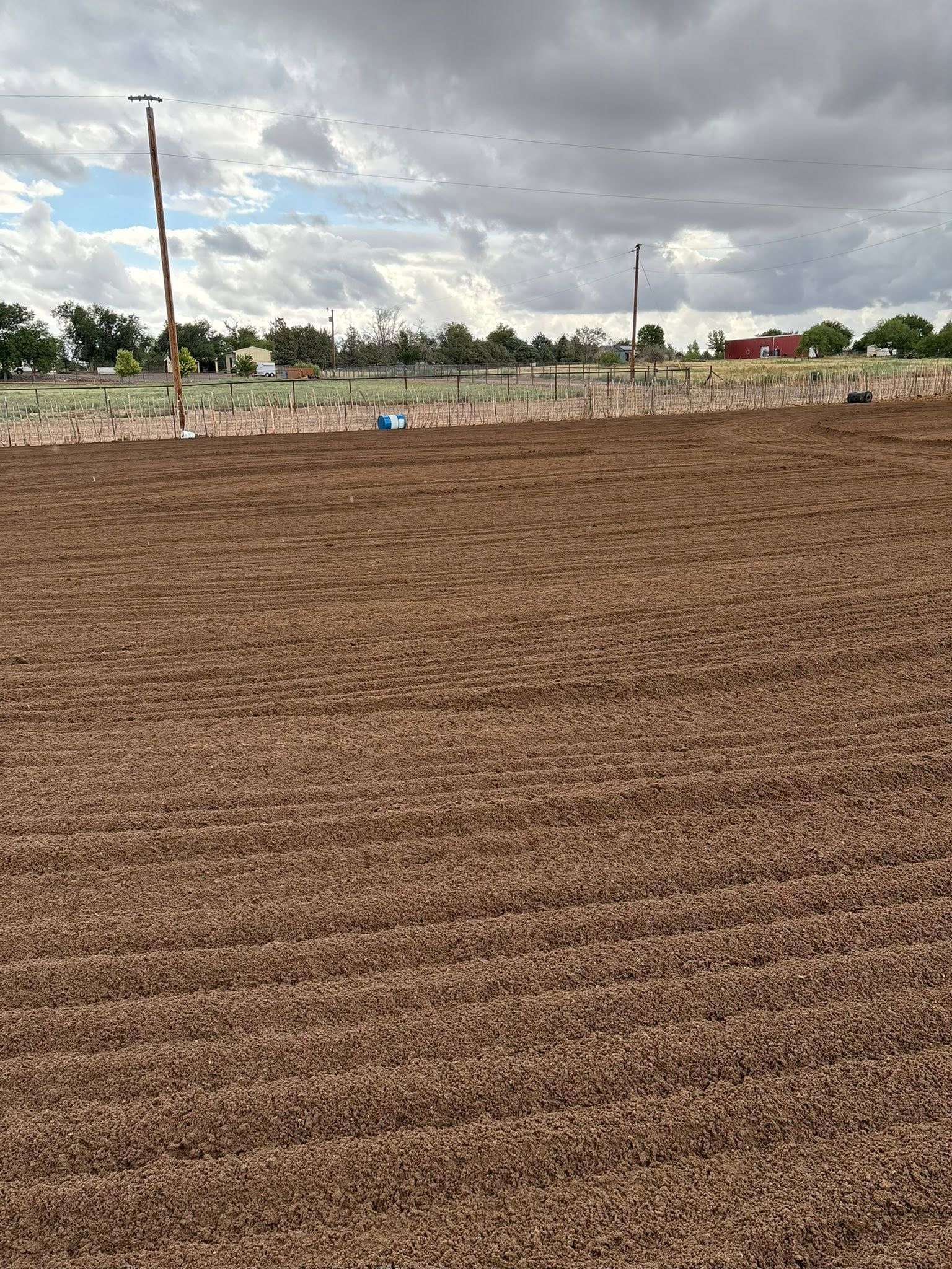Plowed brown field under a cloudy sky, with a fence and distant structures on the horizon.
