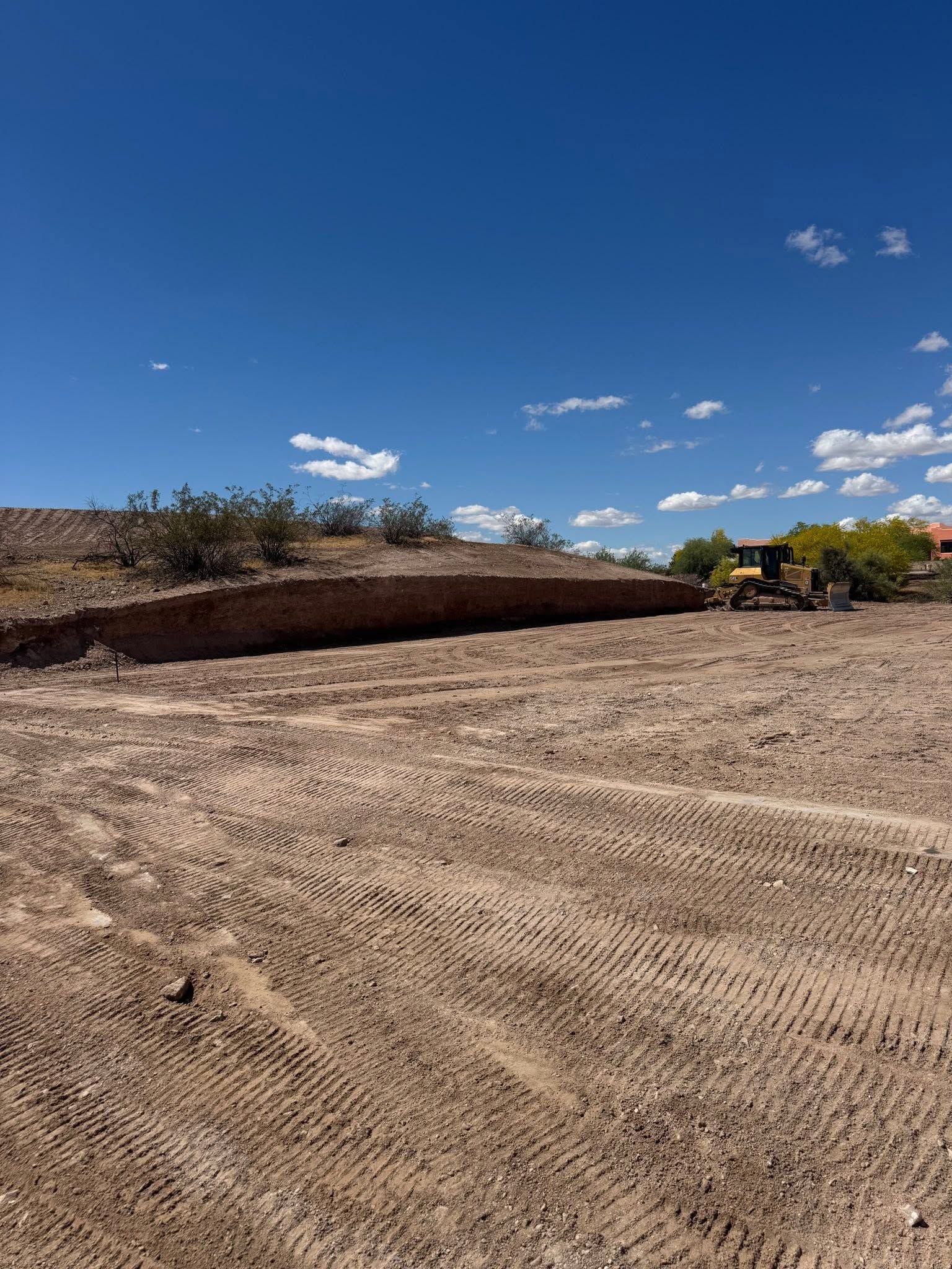 Dirt lot under a bright blue sky, with a low earthen embankment and sparse shrubs in the distance.