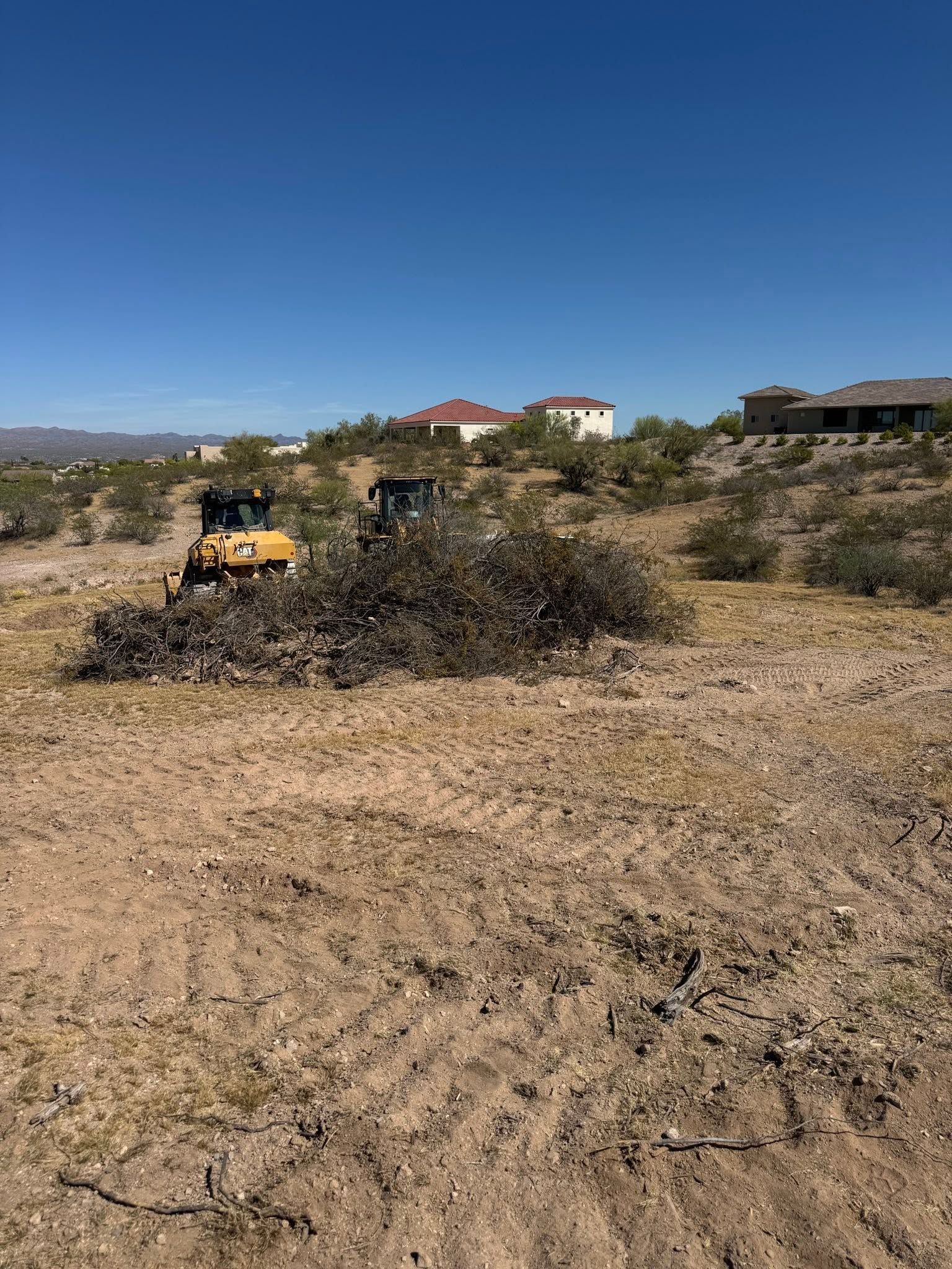 Bulldozer and dump truck moving brush in a dry field with houses in the distance under a clear blue sky