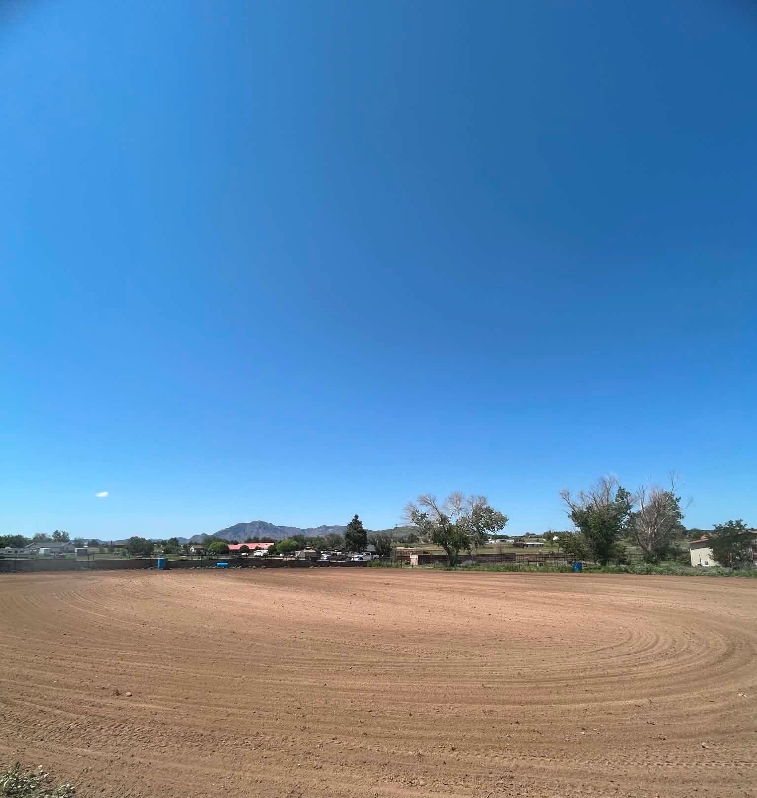 Wide dry dirt field under a clear blue sky with distant trees and buildings on the horizon