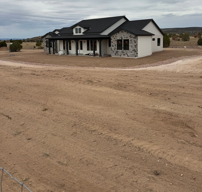Single-story house with stone siding and dark roof on a bare dirt lot in a rural area