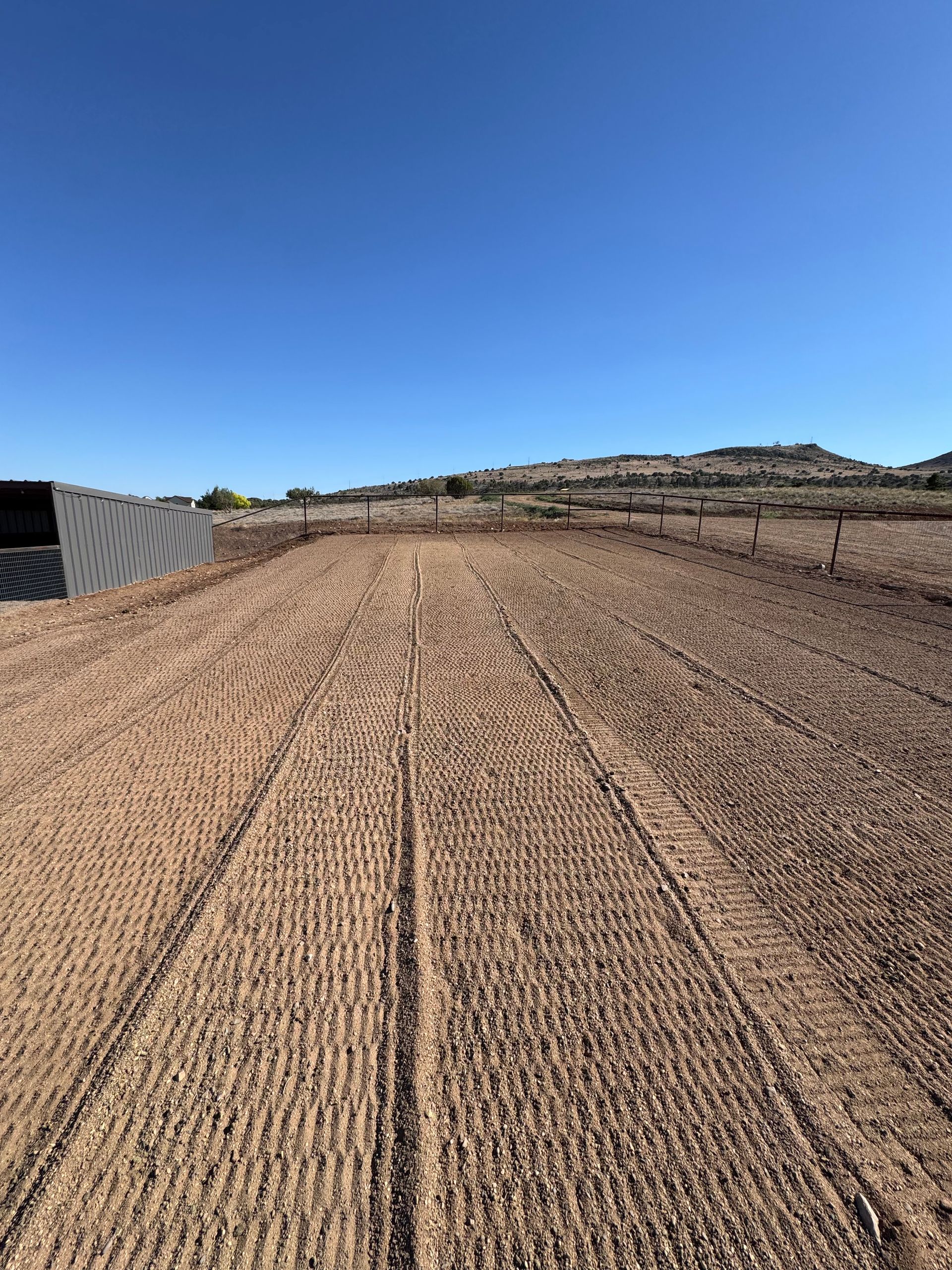 Textured dirt field under clear blue sky, with terraced hills in the distance.