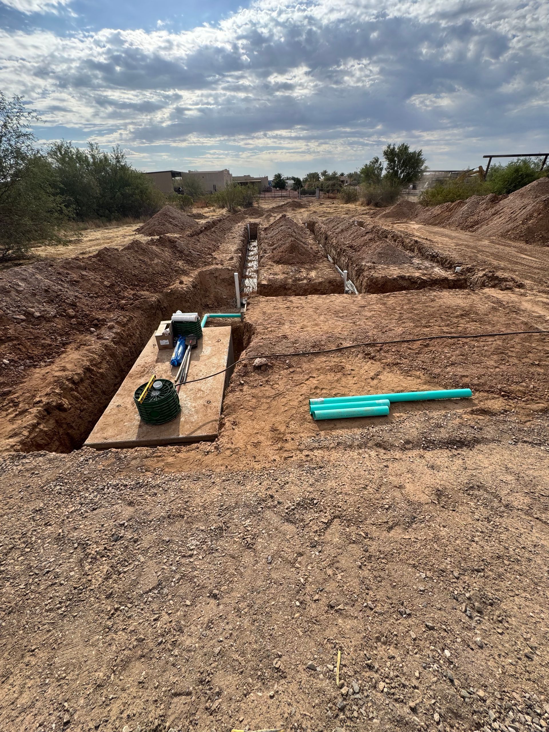 Utility trench in a rocky dirt field with pipes and green equipment under a cloudy sky