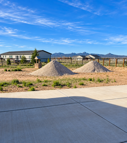Residential lot with two gravel mounds, a fence, houses, and mountains under a blue sky