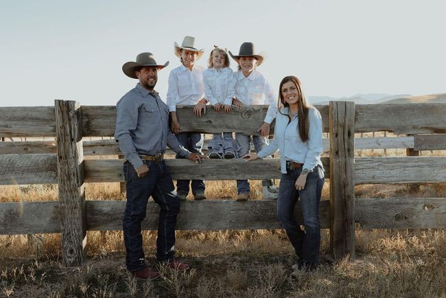 Four people posing by a rustic fence in a dry field, two sitting on the rail and two standing beside them.
