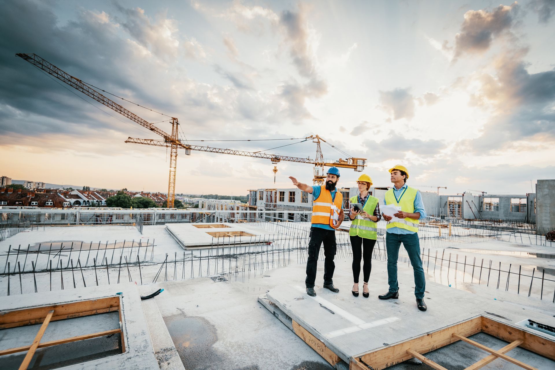 Operai edili in un cantiere, discutono i progetti. Uno indica, altri tengono i documenti. Gru sullo sfondo.