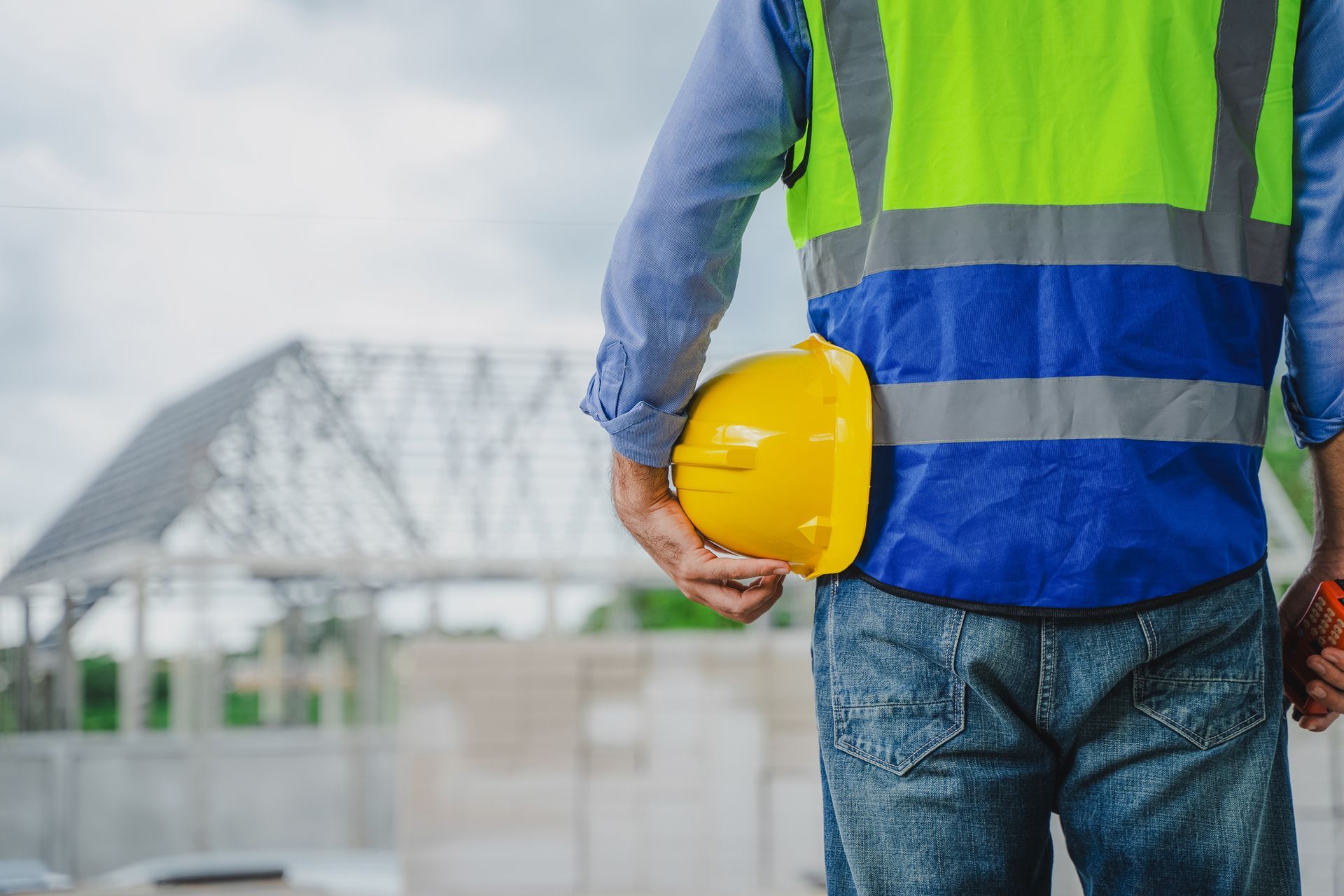 Operaio edile con giubbotto di sicurezza e casco giallo in mano, sullo sfondo un edificio in costruzione.