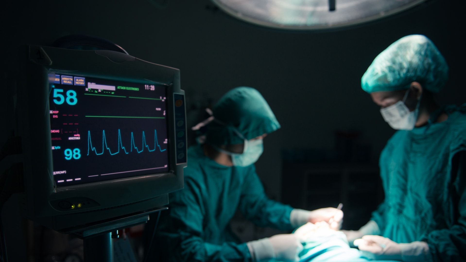Surgical team operating; patient monitor displays vital signs; operating room setting.