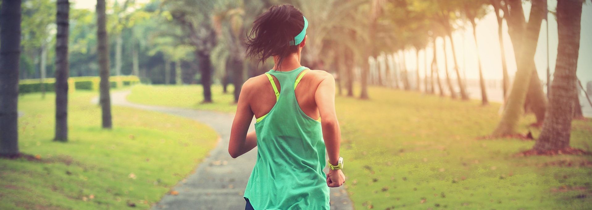 Woman running on a pathway in a park, trees lining the sides. She wears green athletic wear and a blue headband.