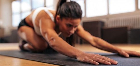 Woman in a white sports bra and shorts stretches on a yoga mat with arms extended forward in a gym.