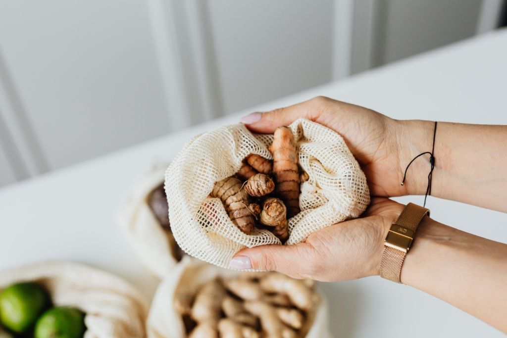 Hands holding a mesh bag filled with ginger and turmeric roots; white table with more produce.