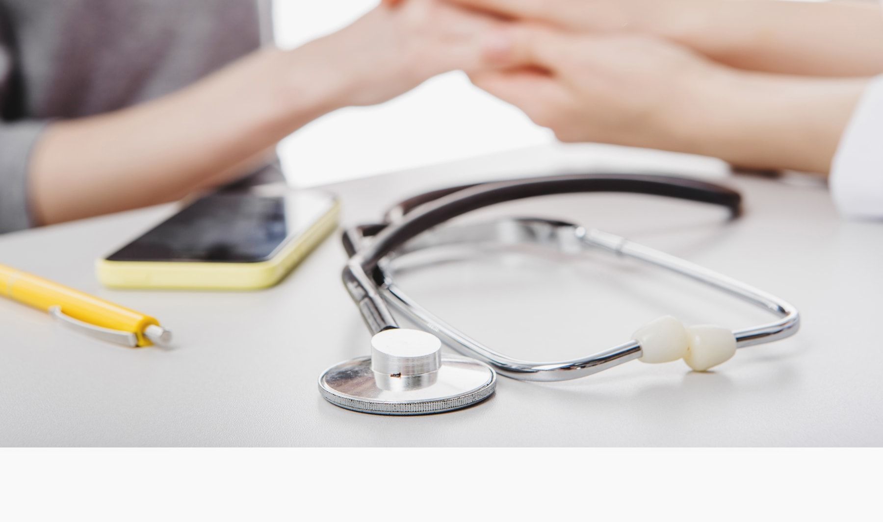 Doctor holding a patient's hand with stethoscope, phone, and pen on a white desk.