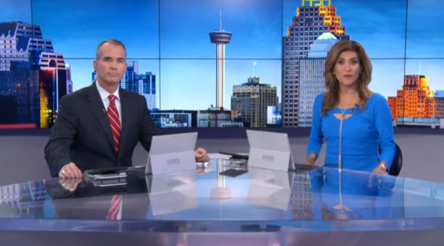 News anchors at desk, cityscape background with tower, one in blue, one in suit.