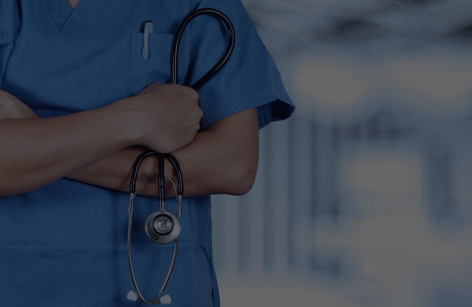 Person in blue scrubs holding a stethoscope, arms crossed, in a blurred hallway.