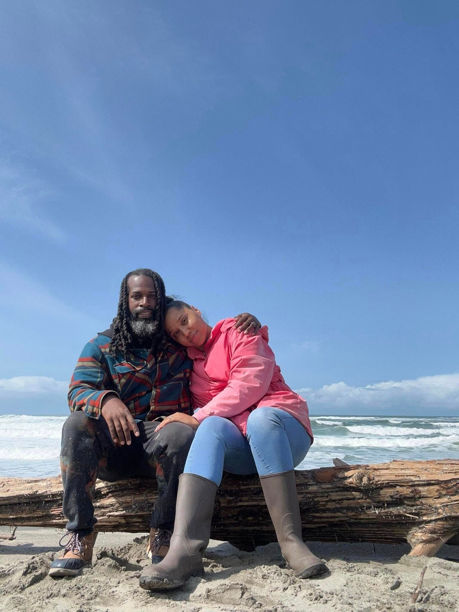 Couple sitting on driftwood at beach, woman resting head on man's shoulder. Blue sky, ocean waves.