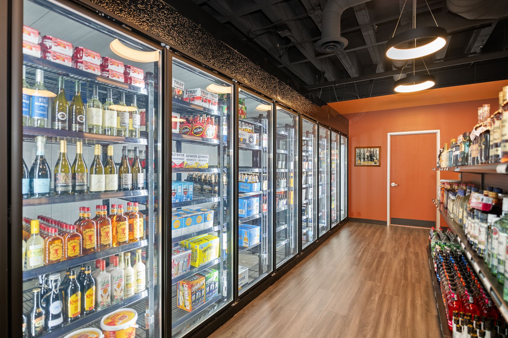 A liquor store aisle with refrigerated glass doors filled with bottles and cans. Wooden floor, orange walls, and black ceiling.