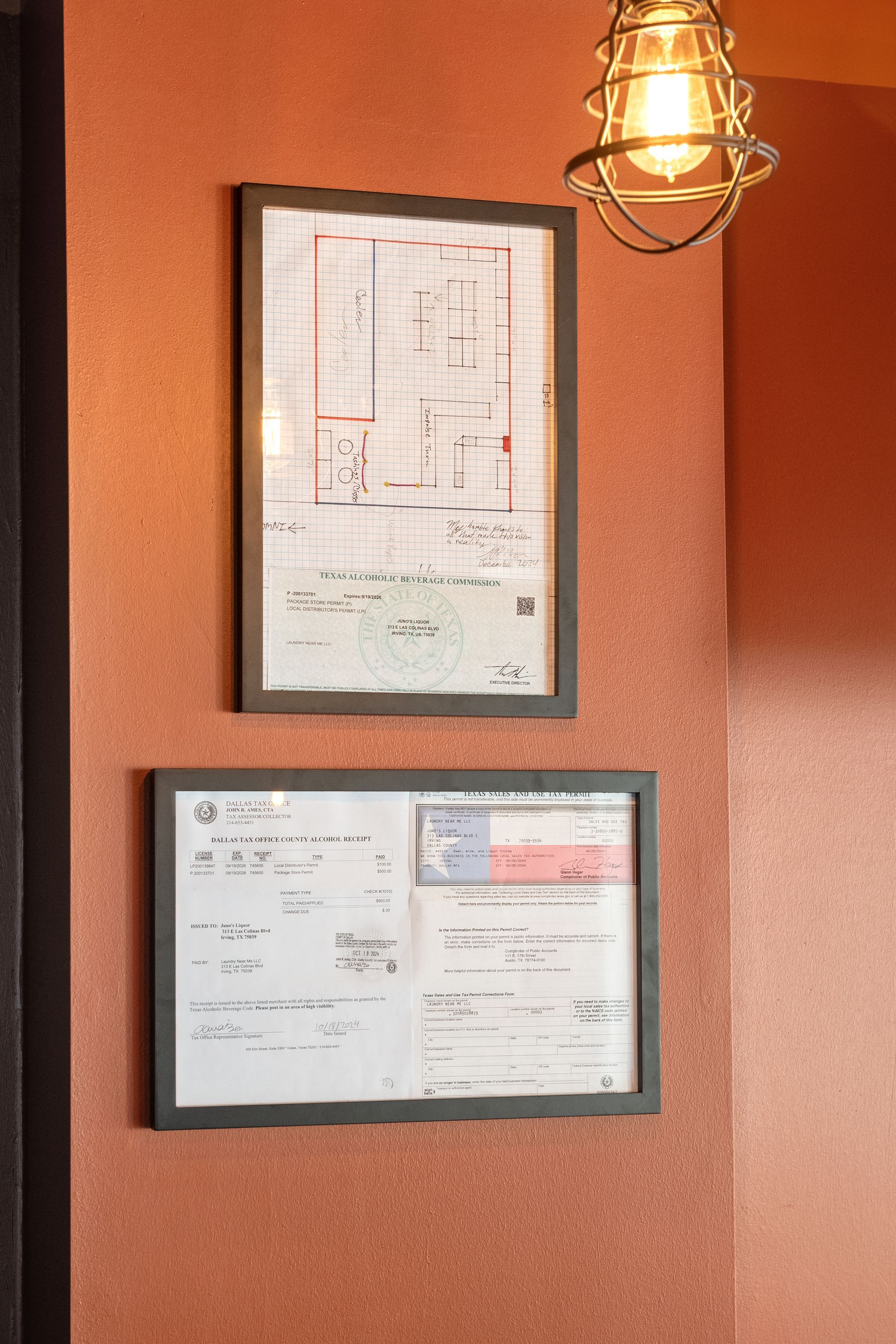 Two framed documents hang on a textured, reddish-orange wall, illuminated by a hanging light.
