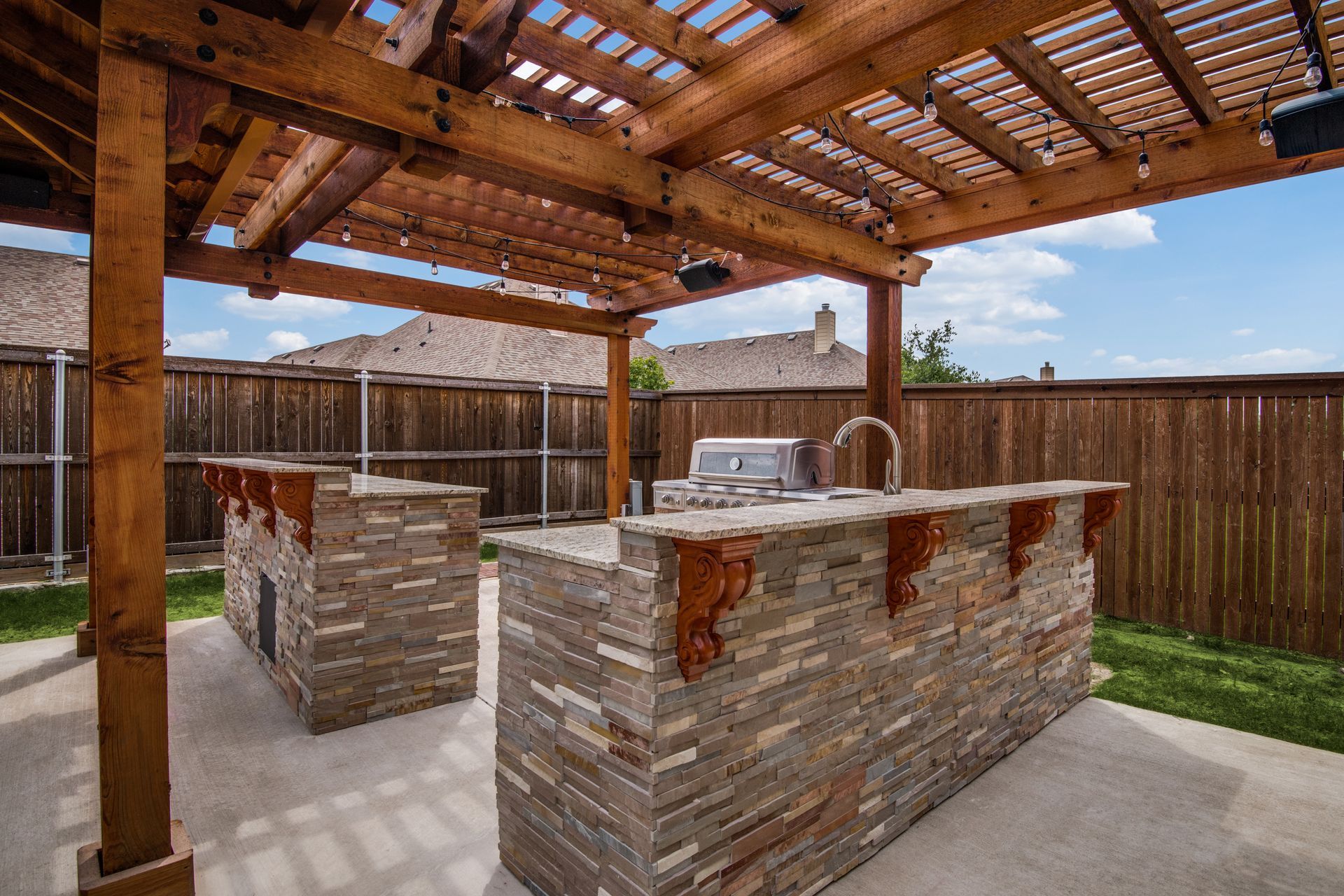 A patio remodel with a wooden pergola and a stone island with a grill underneath it.