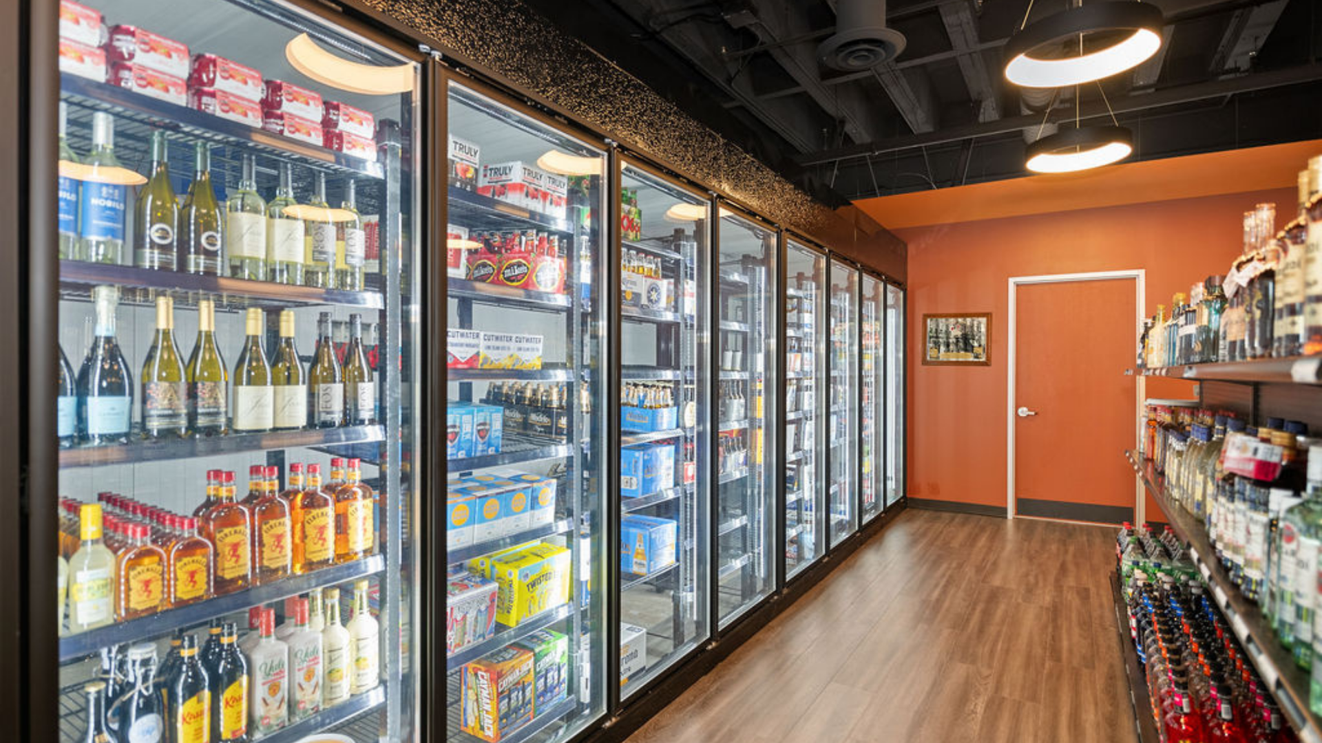 Liquor store aisle with glass-door coolers filled with beverages, wooden floor, orange wall, and black ceiling.