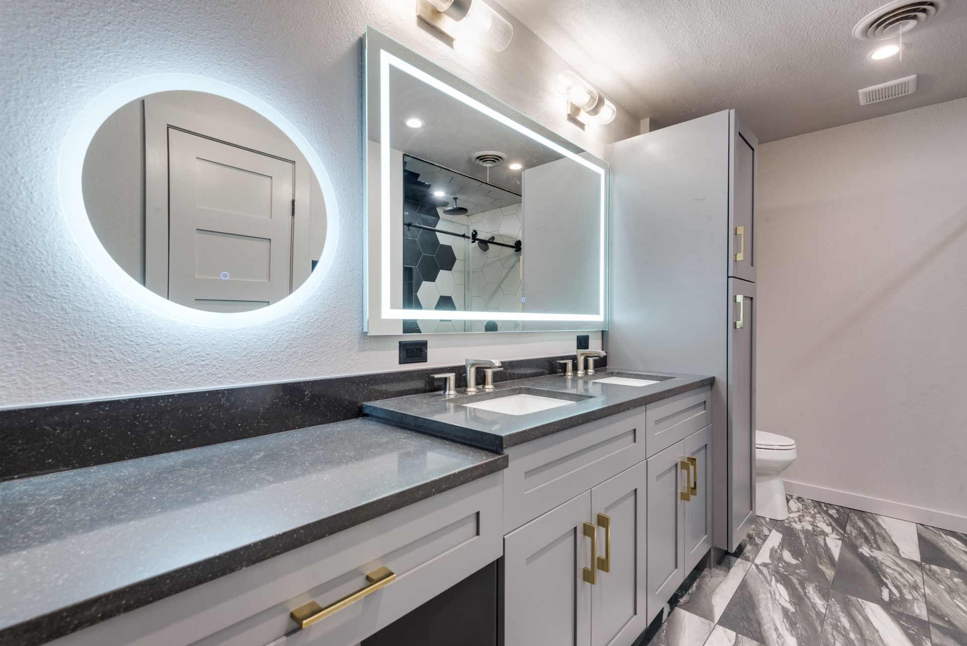 A bathroom remodel with two sinks , a toilet , and a round mirror.