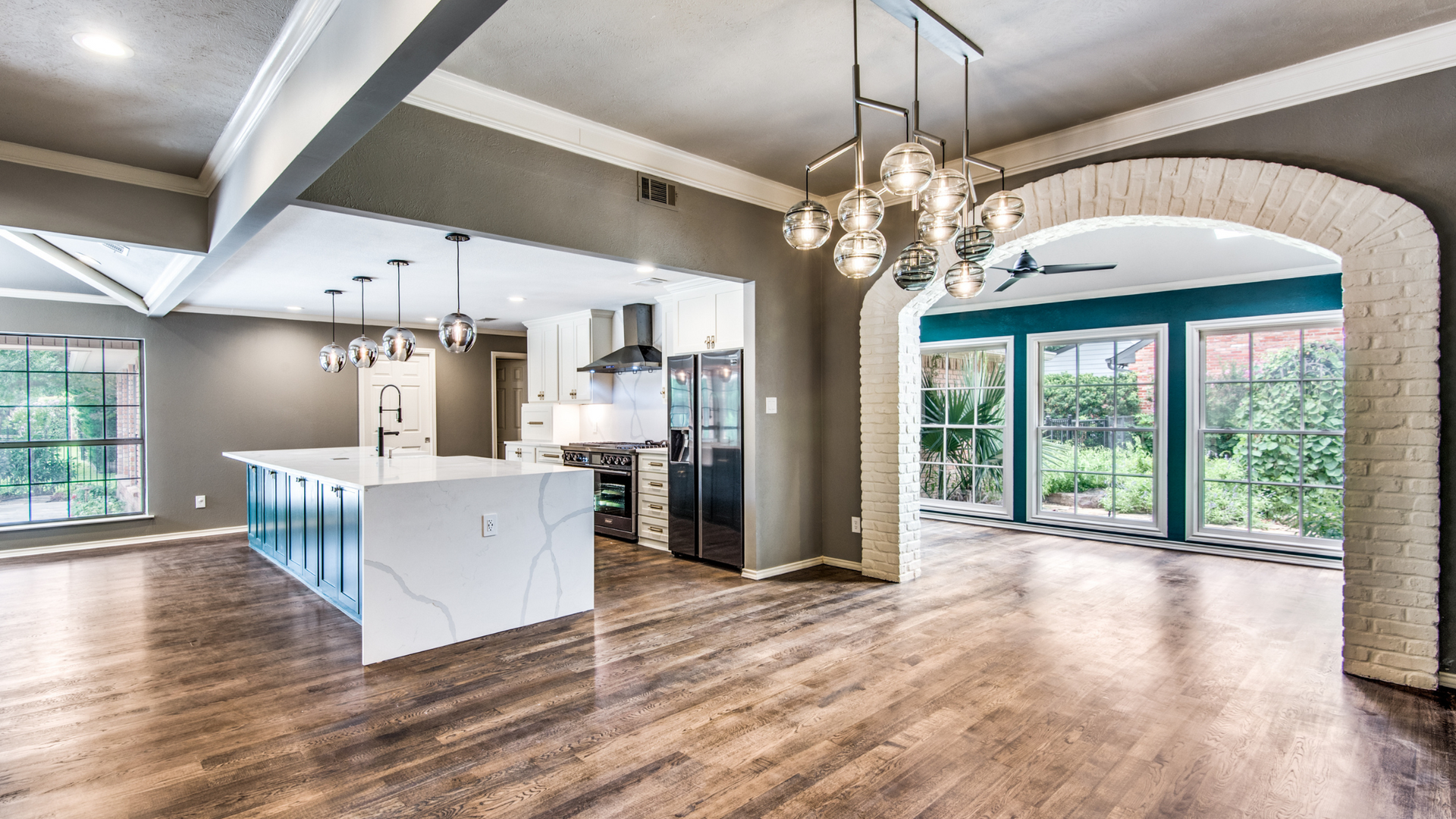 Open-concept kitchen with white island, dark cabinets, and arched doorway leading to a sunroom with greenery.