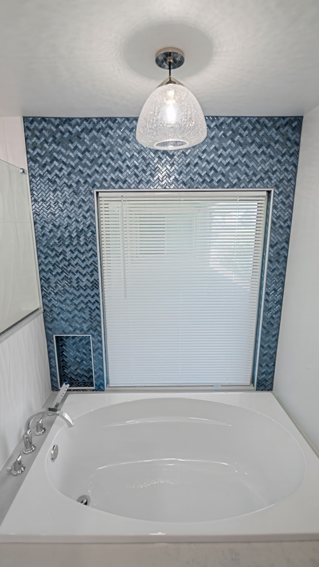 Bathroom with a white tub, blue tile wall, window with blinds, and a hanging light.