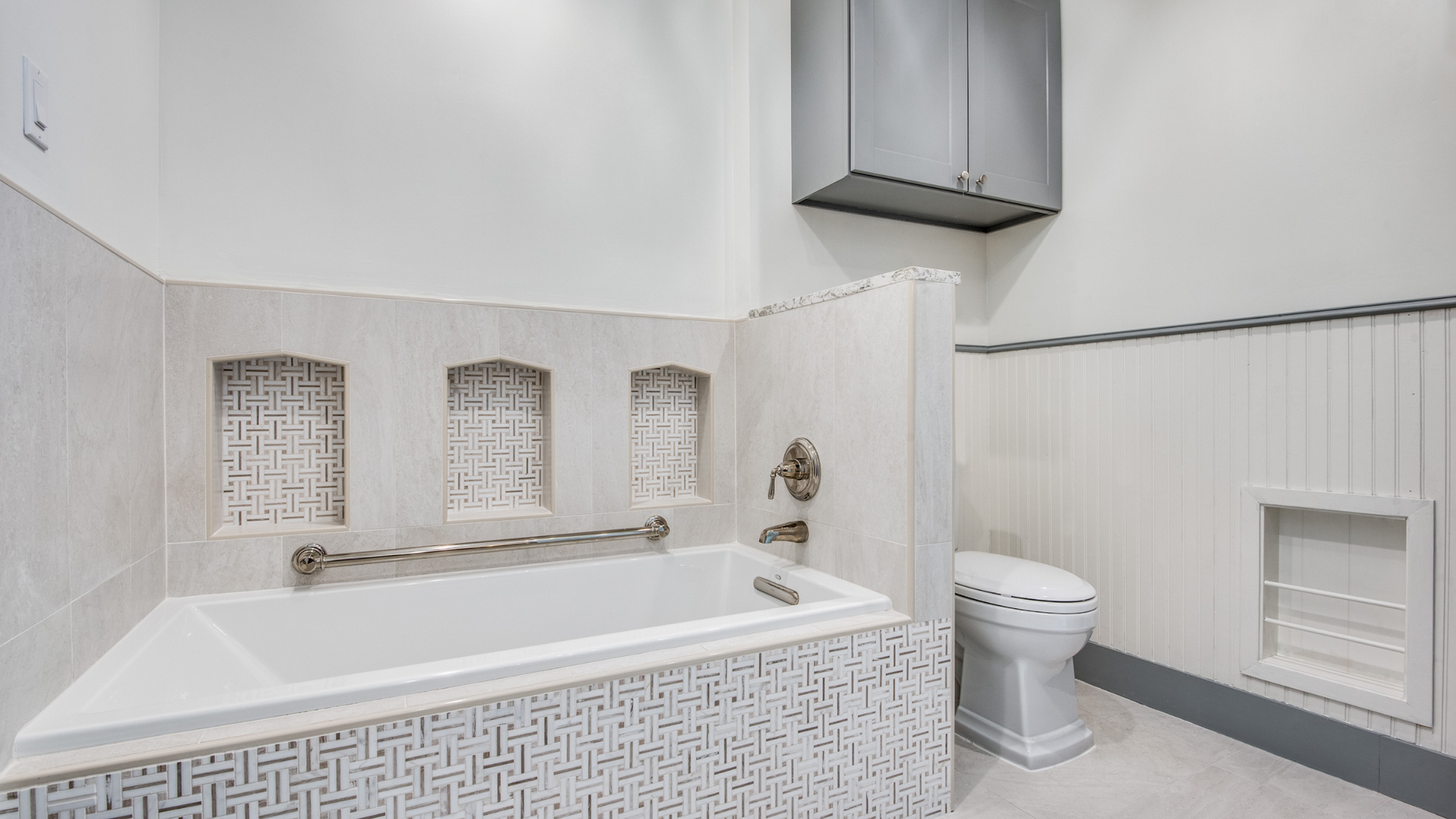 Bathroom with tub, toilet, and gray cabinets. White tile with decorative mosaic.
