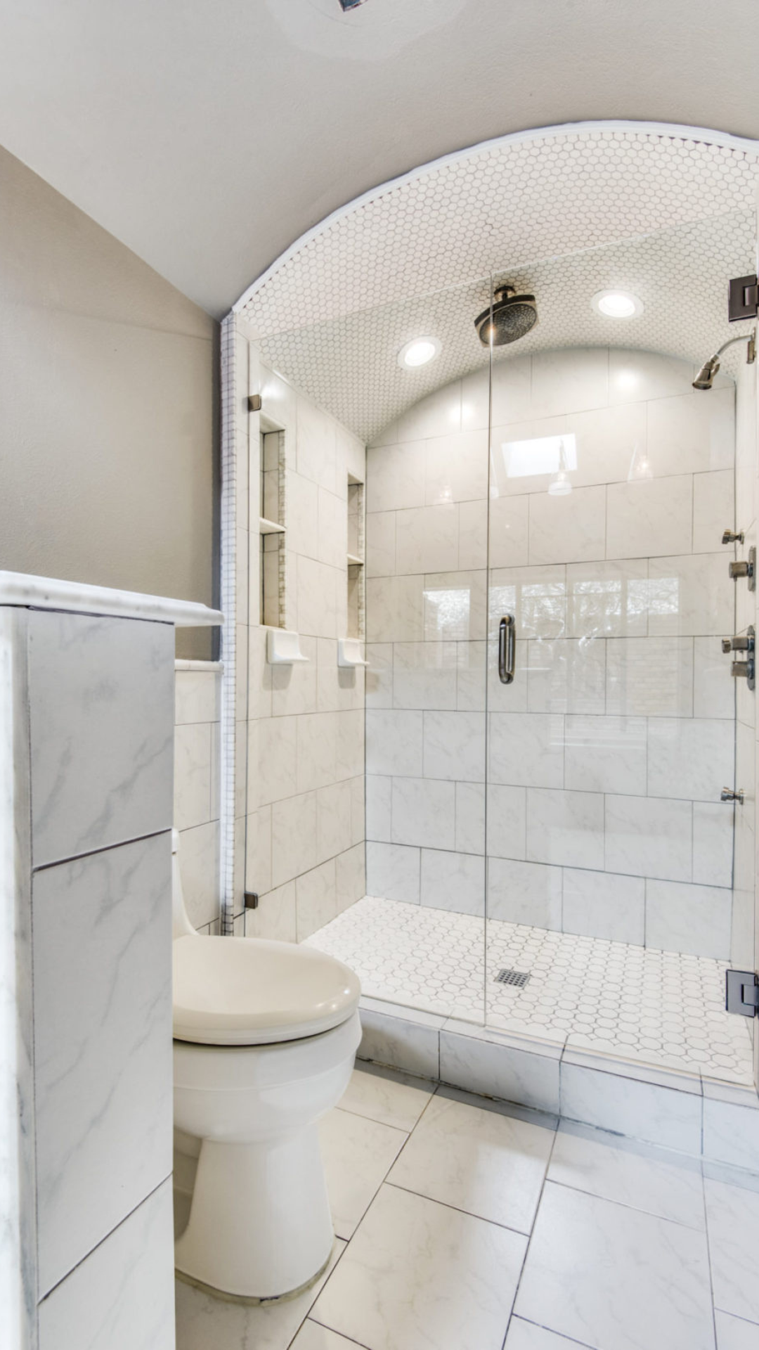 A white bathroom with a toilet, glass shower, and marble-like tiles.