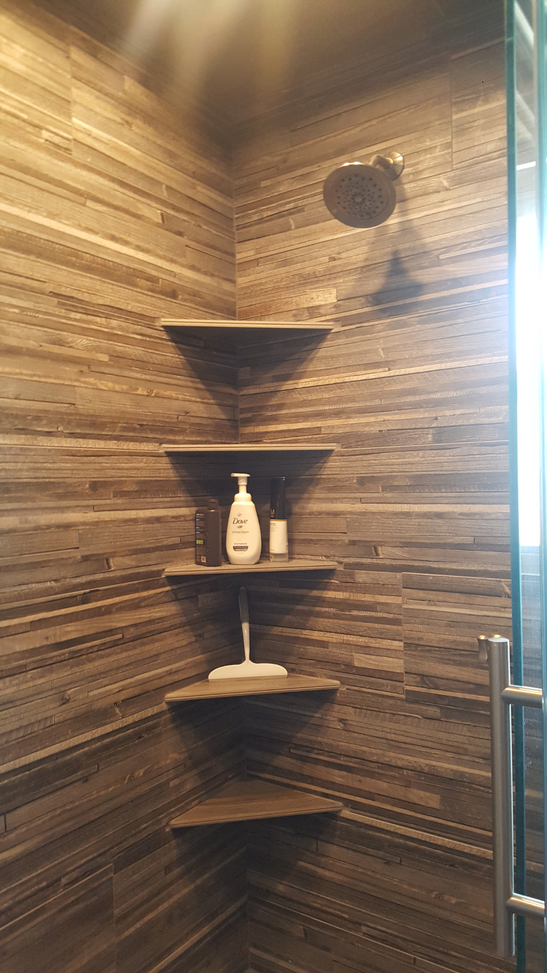 Corner shower with wooden shelves holding toiletries; brown tiled walls.