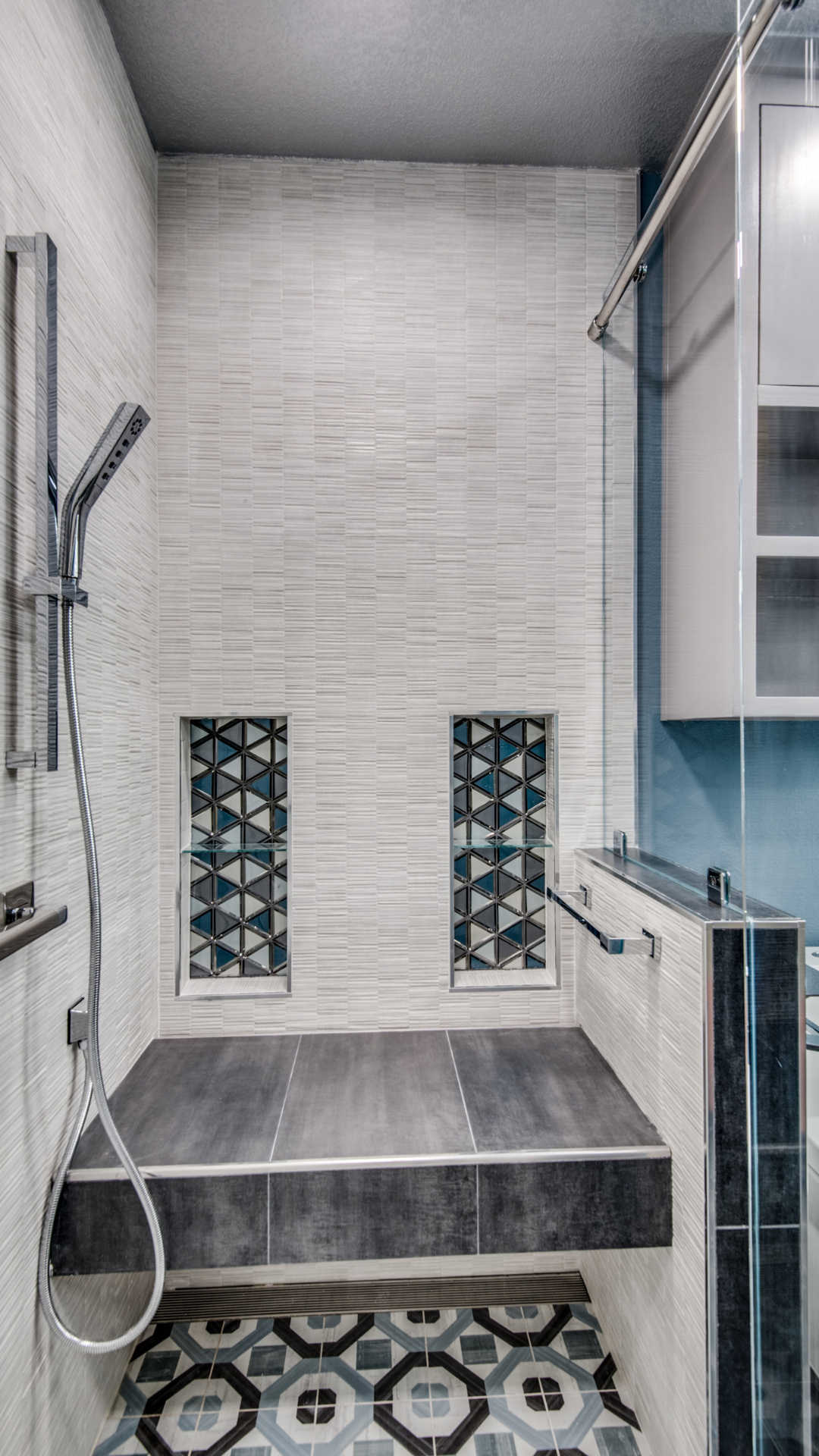Shower with textured white tile, two recessed shelves with patterned glass, gray platform, and patterned floor tile.