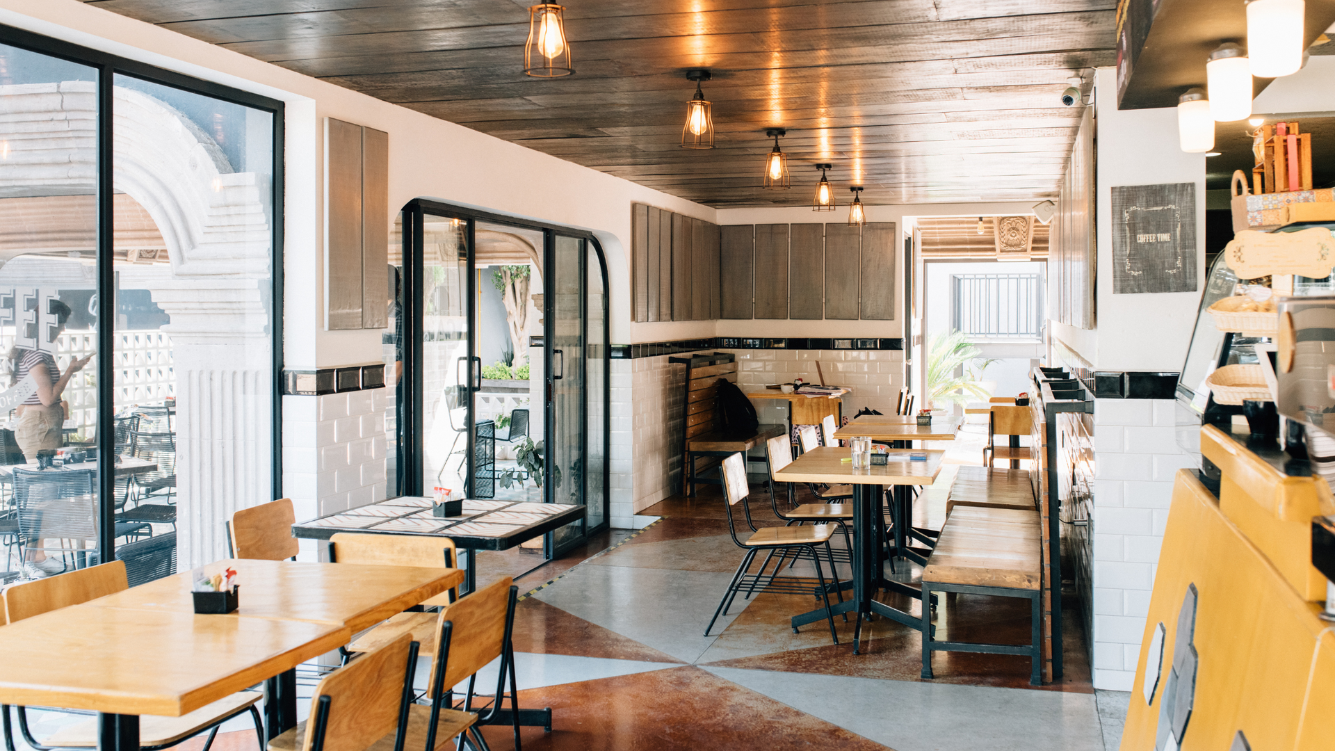 Interior of a cafe with tables, chairs, and large windows. Wooden floors and light fixtures.
