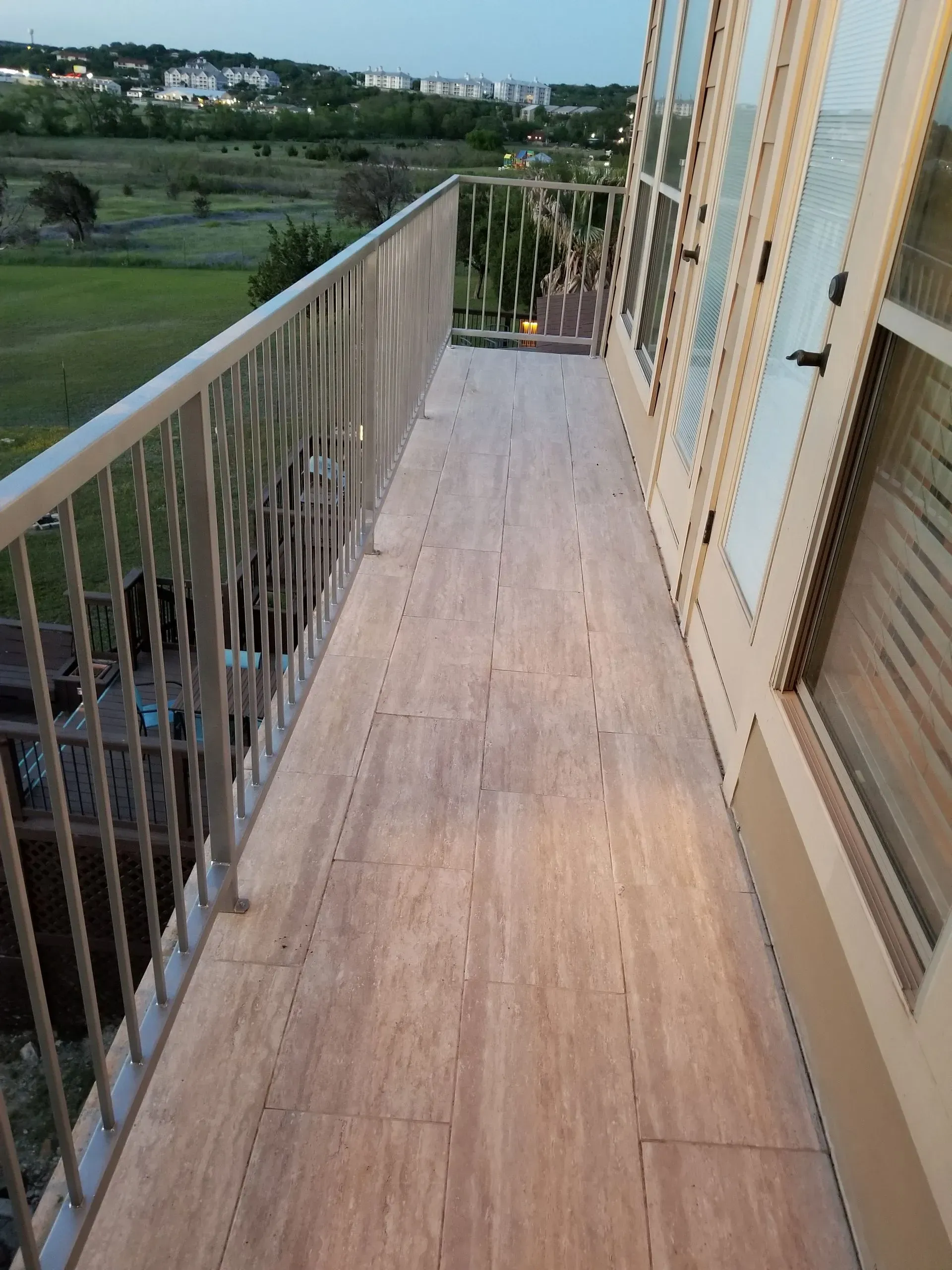 Balcony with wood flooring and metal railing, overlooking a green field and distant buildings.