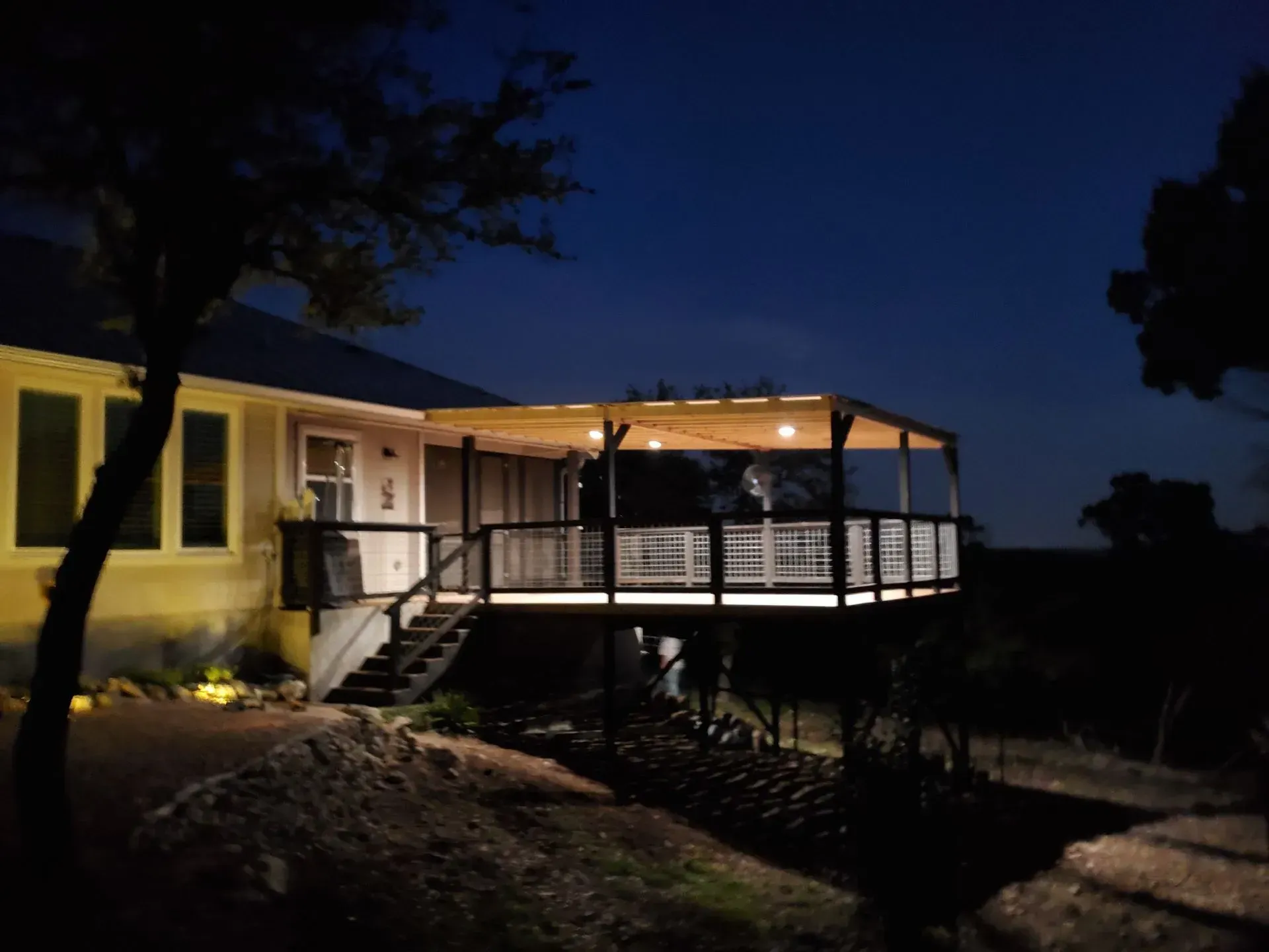 Night view of a house with a lit wooden deck; dark sky, trees, and ground surround.