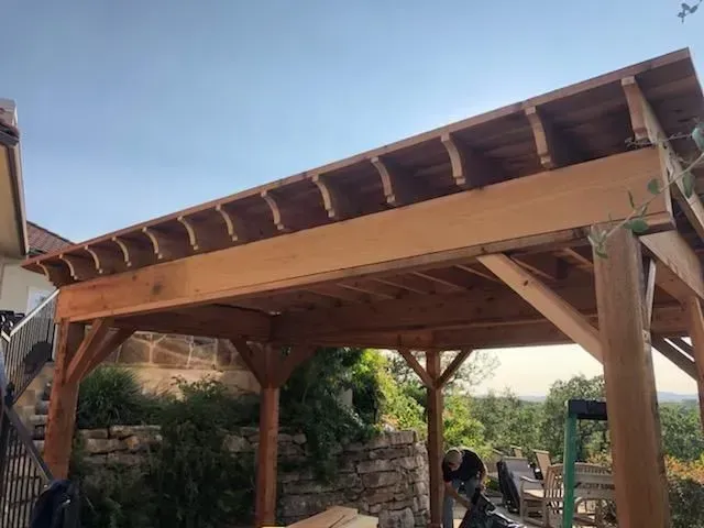 Wooden pergola with a brown roof and supports, overlooking a sunny outdoor scene.