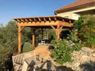 Wooden pergola attached to a light-colored house, built on a sloped yard with greenery.