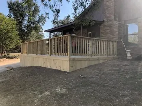 Wooden deck attached to a house with a brick chimney. Wooden railing surrounds the deck, and a dirt yard is in front.