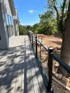 Deck with dark railing and wire cables, extending along a building's side, with trees and a bright sky in the background.