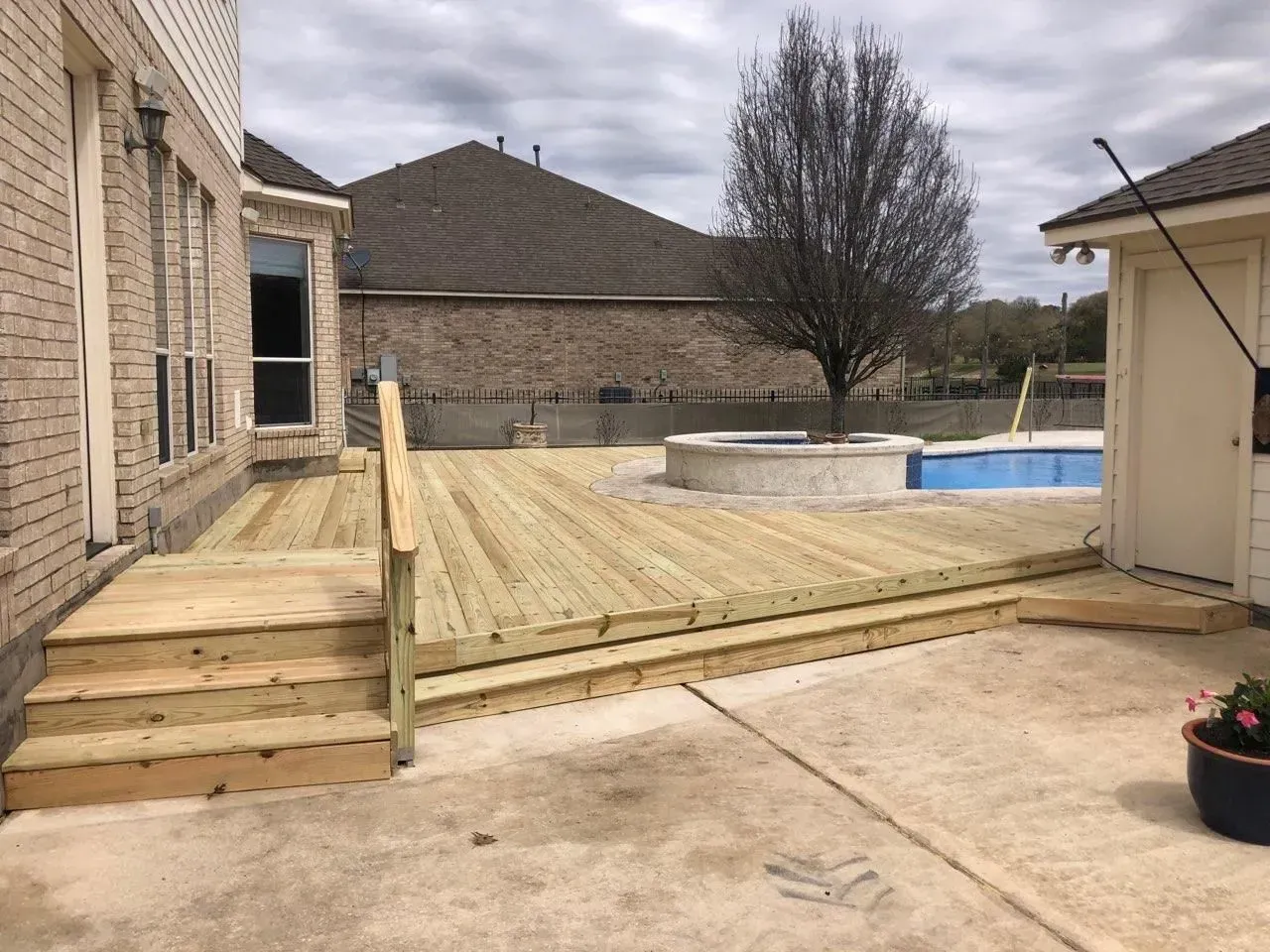 Wooden deck with stairs next to a house and concrete patio, pool in background.